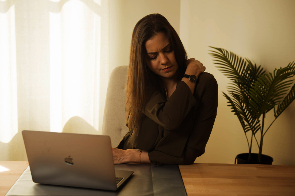 Femme assise au bureau devant l'ordinateur se tient l'épaule et la nuque, son visage est crispé et elle semble avoir une douleur