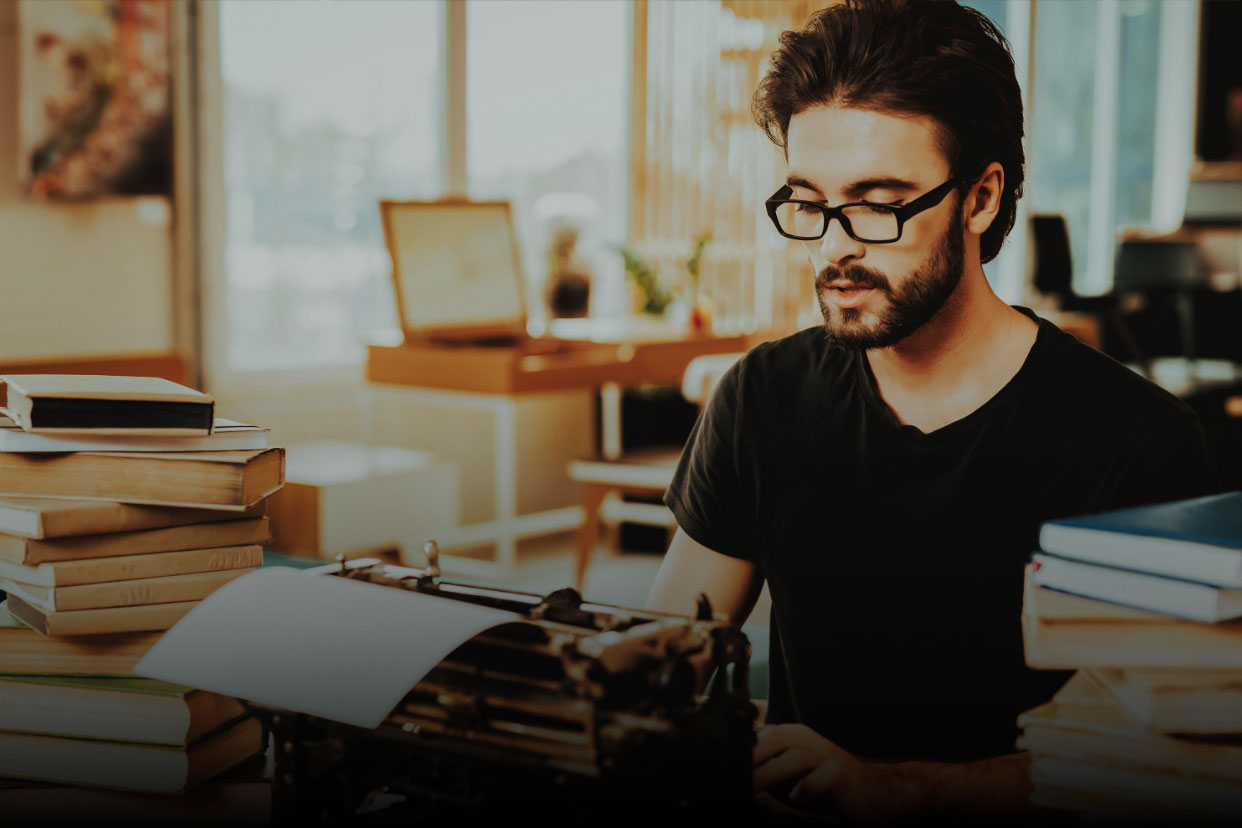 Un homme avec de la barbe et des lunettes est assis à un bureau avec des piles de livres et une machine à écrire. Il est entrain de rédiger un article